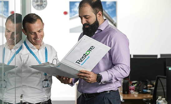A man holds a restore datashred folder and there is another man stood next to him smiling with a lanyard, behind them is a computer