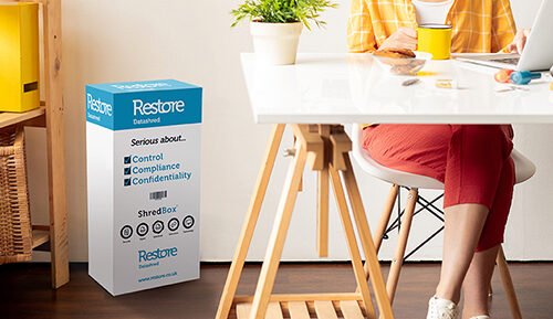 Woman sat at a desk with a blue and white tall shred box on the floor near her